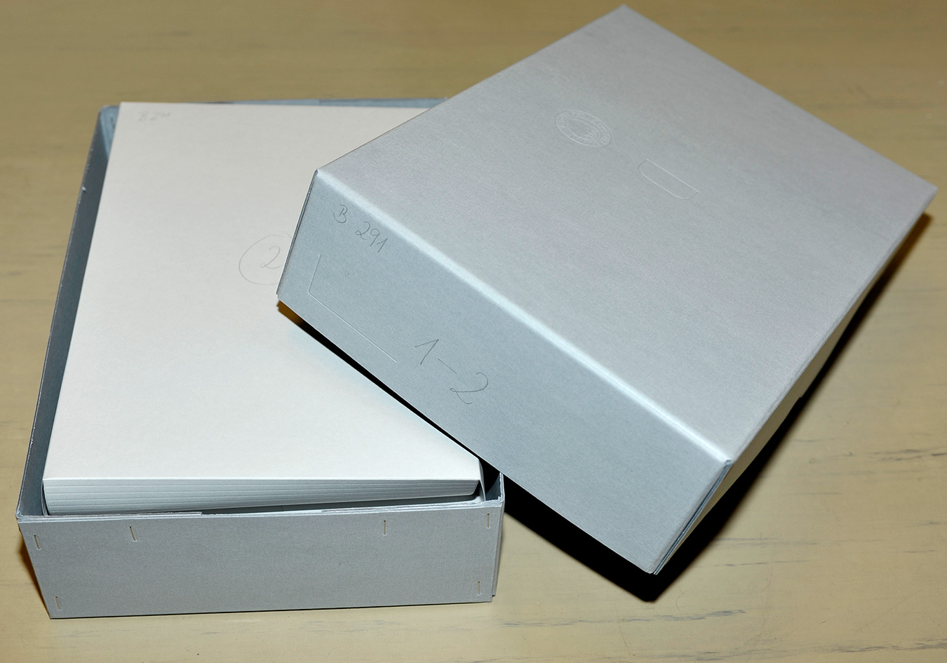 An open gray archival document box with a stack of paper folders inside, resting on a pale wooden table.
